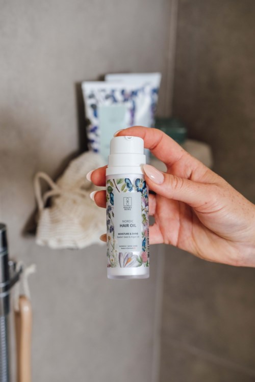 Eine Hand hält eine kleine weiße Flasche mit dem Titel 'HAIR OIL' in einem Raum mit Regalen und Gegenständen im Hintergrund.