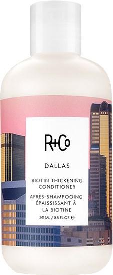 Een witte fles met een stadssilhouettemerk, roze band en zwarte dop, bevat biotine-verstevigende conditioner.