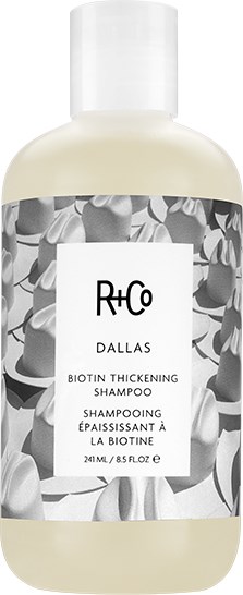 Een witte fles R+Co Dallas Biotin Thickening Shampoo met een zwart-wit bloemenetiket.