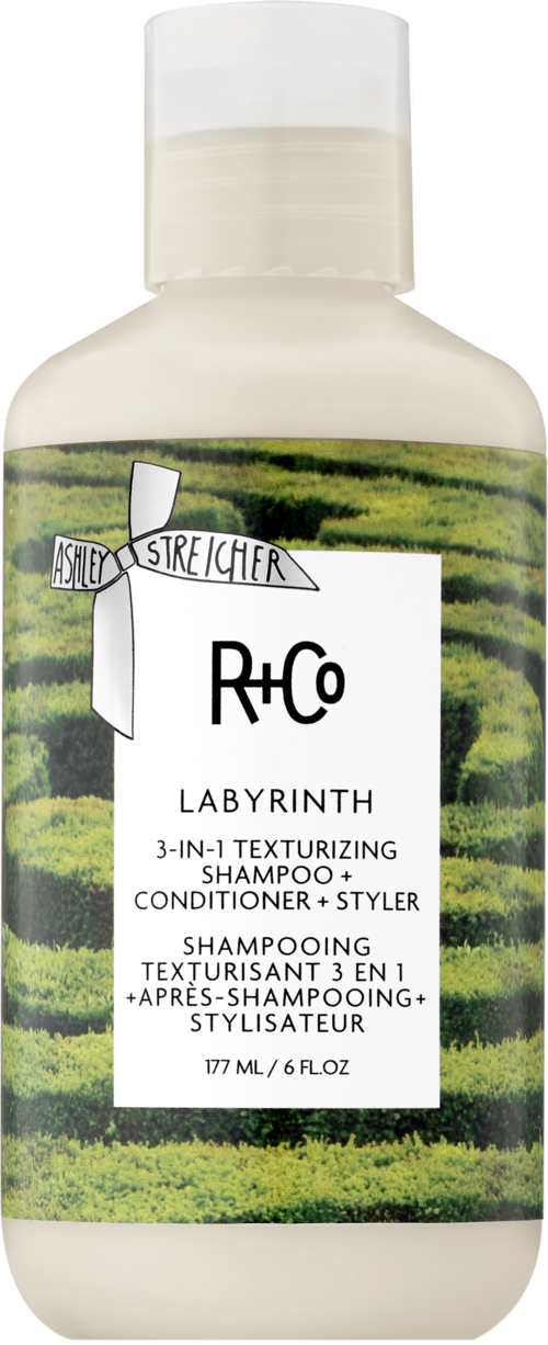 Een witte fles met een groene landschap achtergrond, gemarkeerd R+Co Labyrinth shampoo, conditioner en styler in één.