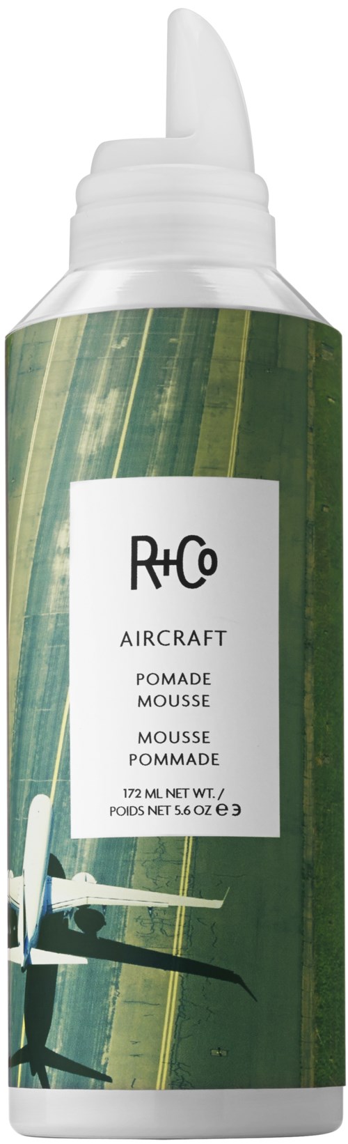 En grønn sprayflaske med hvit tut, merket R+Co, er designet for hårmousse.