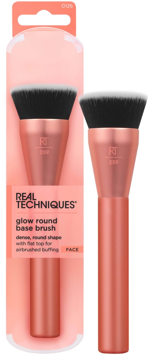 A makeup brush with a pink handle and black bristles, in packaging labeled 'Real Techniques glow round base brush.'