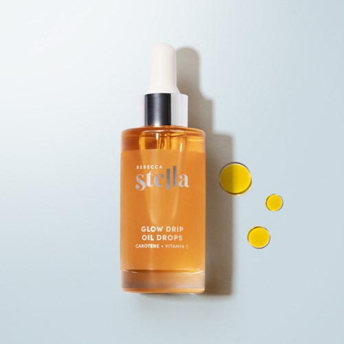 A bottle of Rebecca Stella Glow Drip Oil Drops with yellow capsules beside it, on a light background.