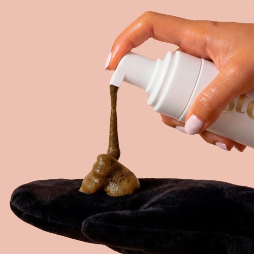 A hand is pouring a brown, foamy substance onto a black surface from a white bottle.