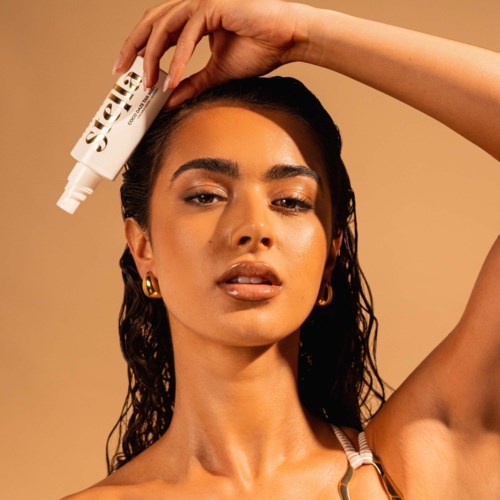 A woman with wet hair and gold earrings holds a skincare product above her head, staring confidently.
