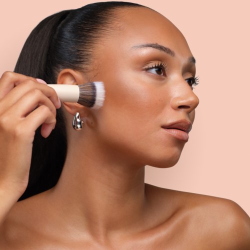 A woman is applying makeup with a brush on her face, looking to the side, with clear skin and long dark hair.