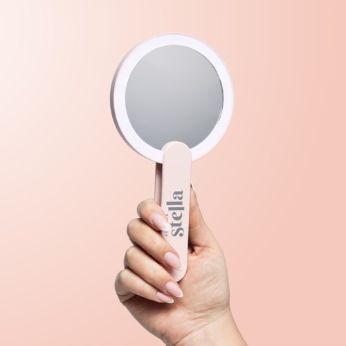 A hand holds a small mirror with the word "Stella" on the handle against a pink background.