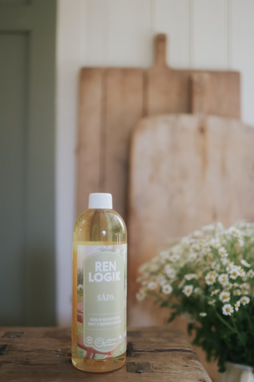 A bottle of cleaning liquid labeled 'REN LOGIK SÄPA' placed on a wooden table, with flowers in a pot nearby and a wooden board in the background.