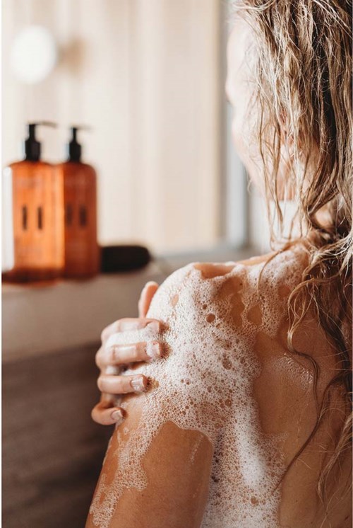 En person med vått, lockigt hår masserar in tvål eller lotion på axeln i ett badrum.