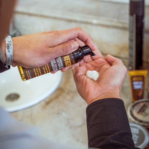 A person applies a grooming product from a bottle onto their hand in a bathroom.