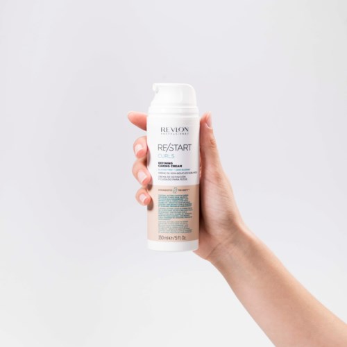 Eine Hand hält eine weiße Flasche mit Revlon-Haarprodukt vor einem schlichten Hintergrund.