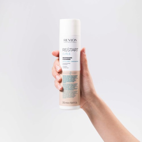Eine Hand hält eine weiße Flasche mit Revlon Haarreiniger vor einem grauen Hintergrund.