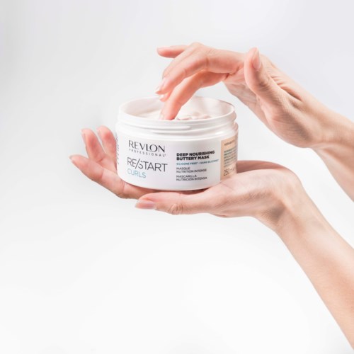 Eine Person verwendet die Finger, um Creme aus einem weißen Behälter mit dem Revlon-Haarmaske-Label zu entnehmen.