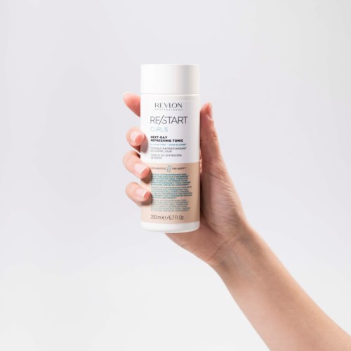 Eine Hand hält eine weiße Flasche mit blauer Schrift für Revlon Re/Start Haar-Tonika. Einfacher Hintergrund.