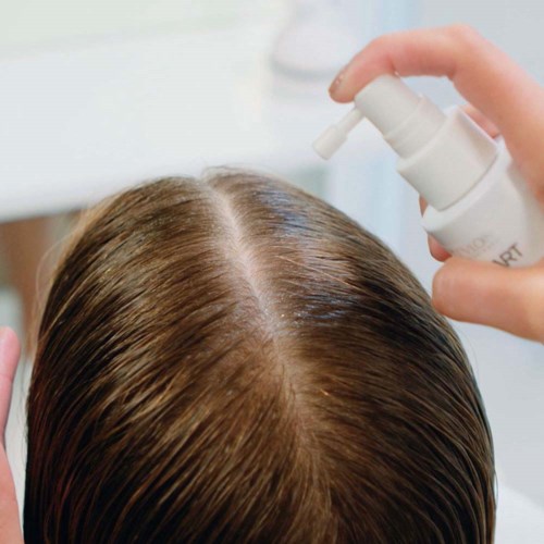 Eine Person sprüht Haarpflegeprodukt auf die Kopfhaut, fokus auf braune Haare.