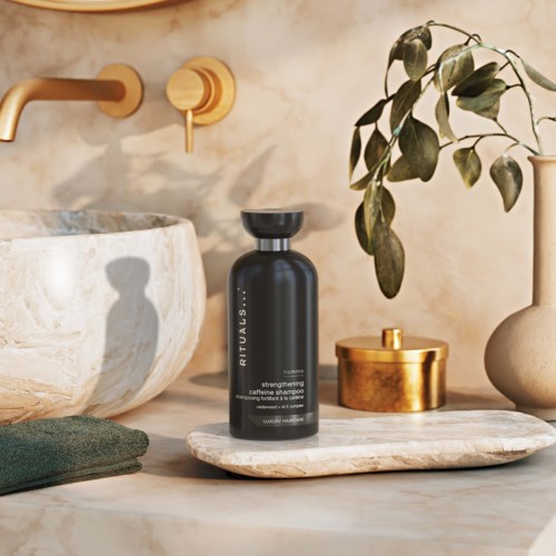 Eine schwarze Flasche Shampoo auf einem Marmorrahmen neben einer grünen Pflanze, einer goldenen Dose und einem Waschbecken.