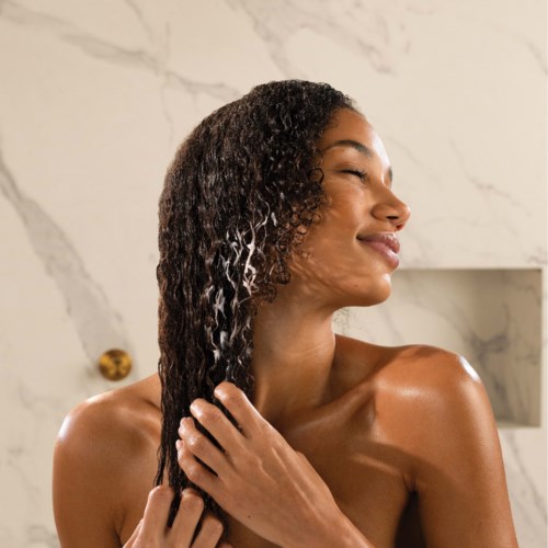 A woman with textured, wet hair is smiling with eyes closed in a marble-tiled shower.