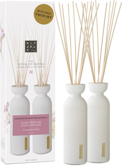 Två vita aromdiffusorer med långa pinnar i vita flaskor, förpackade med etikett och dekorativa element.