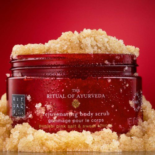 Ein roter Behälter mit dem Titel The Ritual of Ayurveda, Körperpeeling und Sand darum auf rotem Hintergrund.