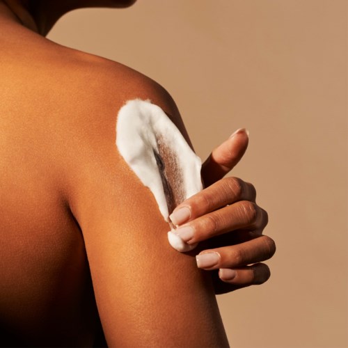 A person applies white cream or lotion on their arm against a plain background.