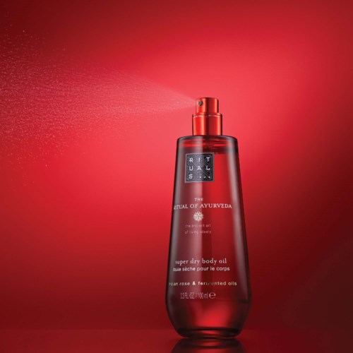 Eine rote Körpersprühflasche mit schwarzem Etikett und Sprühkopf, die feinen Nebel versprüht vor rotem Hintergrund.