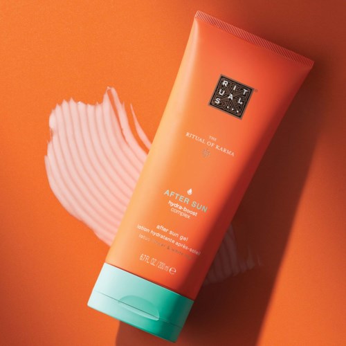 Orangene Tube mit Ritual of Karma After Sun Gel und türkisfarbenem Verschluss auf orangefarbenem Hintergrund. Weißer Pinselstrich im Hintergrund.