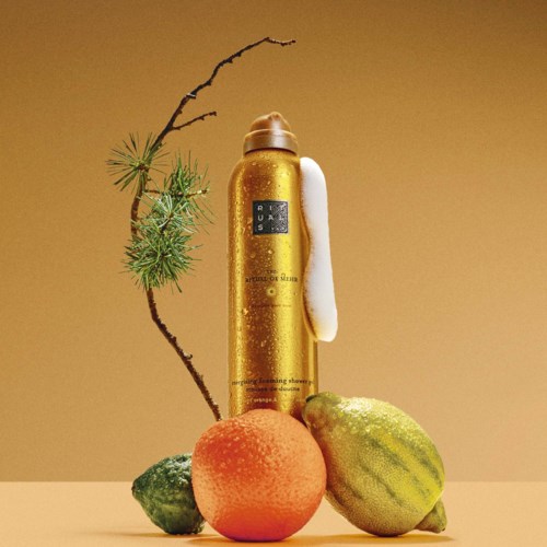 A golden bottle stands behind a peach, a small green fruit, and a striped melon, with a branch and pine needles nearby.