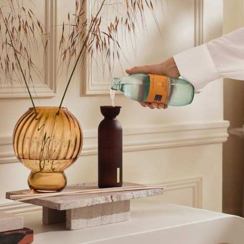 A person pours a bottle of water into a dark vase on a small table with a large round orange vase nearby.