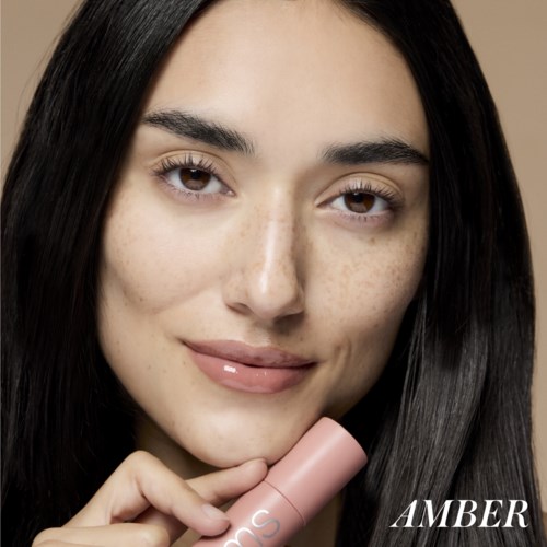 Eine Frau mit langen dunklen Haaren hält eine rosa Flasche, lächelt sanft, mit klarer Haut und natürlichem Make-up.