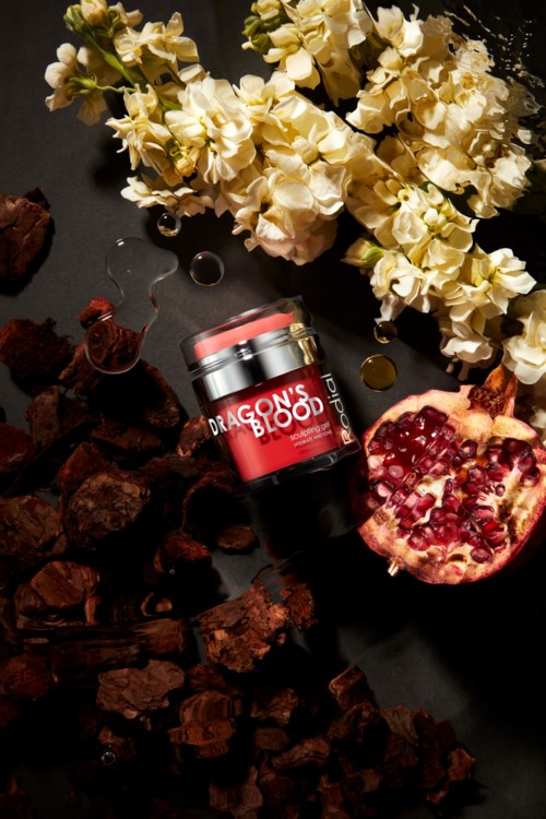 A jar of skincare cream with pomegranate, white flowers, and dark rocks on a black background.