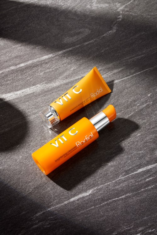 Twee oranje lipbalsemtubes met 'VIT C' op een donkere ondergrond. Licht valt erop, waardoor schaduwen en glans ontstaan.