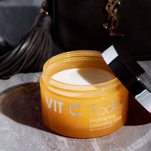 Geel blik met vitamine C-verhelderende reinigingspads, witte pads binnenin en een zwarte deksel. Op de achtergrond een zwarte tas met gouden logo.