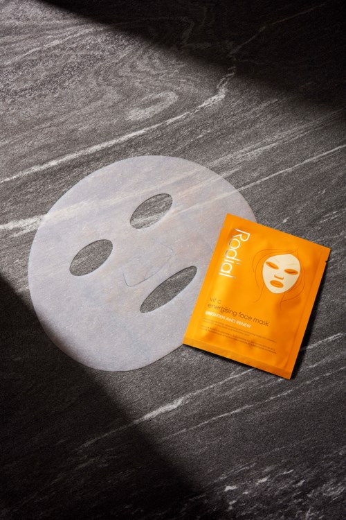 Een gezichtsmasker en een klein oranje zakje op een donkere ondergrond. Het masker heeft oog- en mondgaten, het zakje heeft een gezicht afdruk.