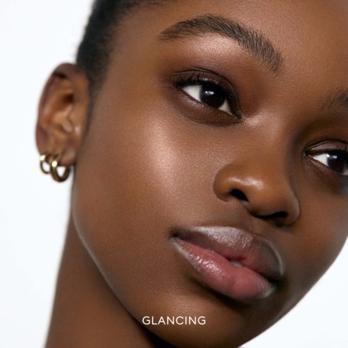 Close-up of a woman's face showing smooth skin, dark eyes, full lips, and small earrings. The word 'GLANCING' appears.