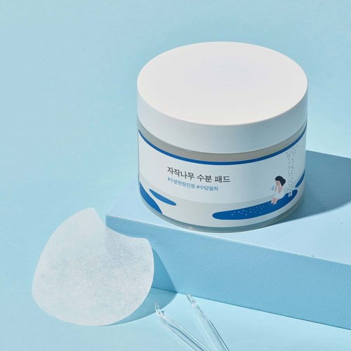 Ein Hautpflegeprodukt in einer runden weißen Dose mit koreanischem Text, neben einem Wattepad auf hellblauem Hintergrund.