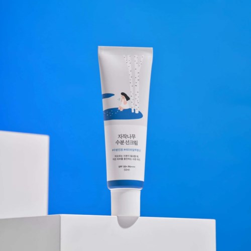 A white tube of skincare cream stands on a white platform against a blue background, showing a girl sitting near water.