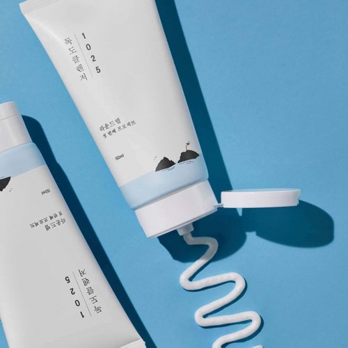 Two white tubes with Korean text, one with a blue cap, on a blue background. One tube has a squiggly line of cream.