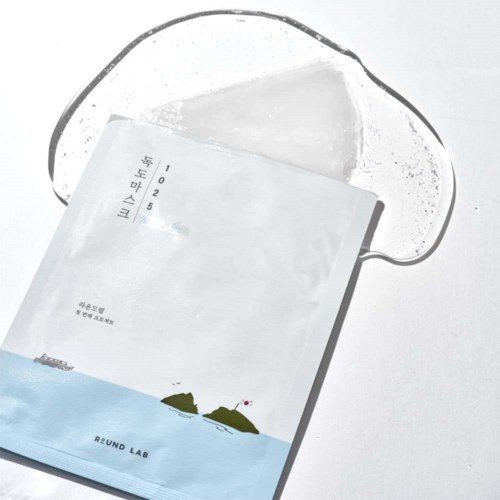 Eine klare Gesichtsmaske liegt über einem weißen Briefumschlag mit kleinen Illustrationen von Hügeln, Wasser und einem Boot.