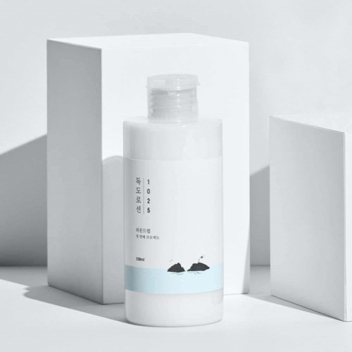 Eine weiße Flasche mit koreanischem Text und kleinen Walfisch-Illustrationen, in einem minimalistischen weißen Raum mit geometrischen Blöcken.