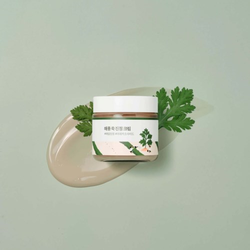 A jar of cream with green leaves, placed on a light green surface with cream spread nearby.