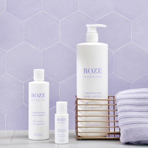 Drei weiße Flaschen mit Shampoo oder Lotion mit Lavendeltext, ein gefaltetes Handtuch und ein Drahtkorb an einer lilafarbenen Fliesenwand.