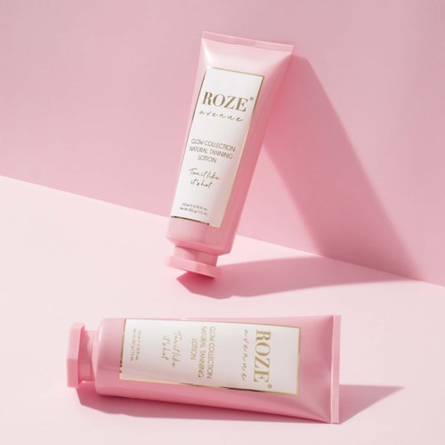 Two pink tubes of skin lotion with gold and white labels, one standing and one lying down, on pink and white background.
