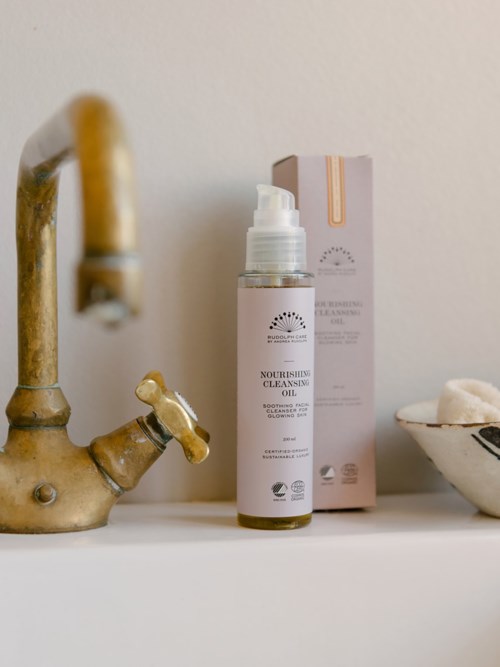 A cleansing oil bottle with a box behind it, a faucet, and a sponge on a white surface.