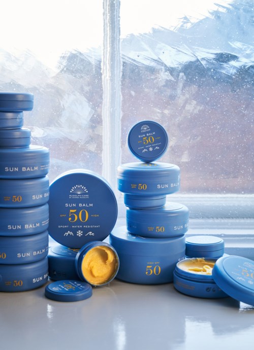 Stacks of blue sun protection tins with SPF 50 are on a windowsill. Some lids are open, showing yellow balm.