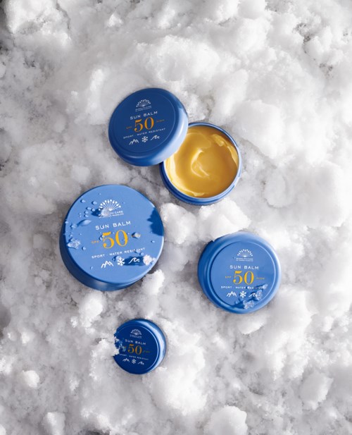 Four blue containers of sun balm with SPF 50 sitting on snow, one open showing yellow balm inside.