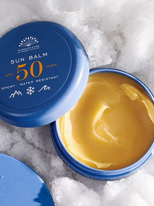 A blue container of sun balm with SPF 50 is open on snow, showing yellow balm inside.