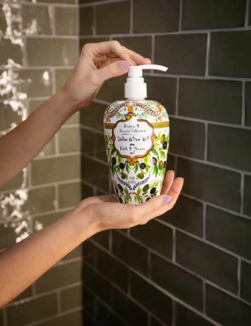 A decorated bottle with a pump, labeled 'Extra Virgin Olive Oil,' held by two hands against a brick wall.