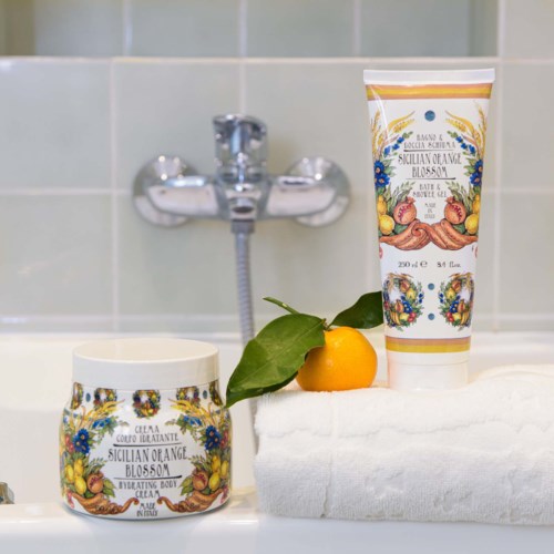 A jar and a tube with colorful floral and fruit designs sit on a white towel, with a small orange and leaf nearby.