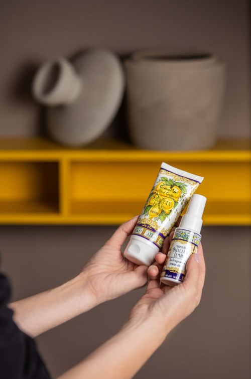 A hand holding two skincare products with colorful labels in front of a yellow shelf and decorative vases.