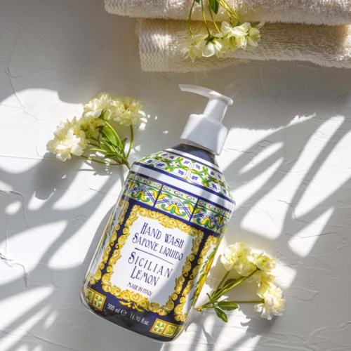 A bottle of liquid hand wash with a Sicily Lemon scent. Surrounded by white flowers and towels, with shadows cast on the surface.